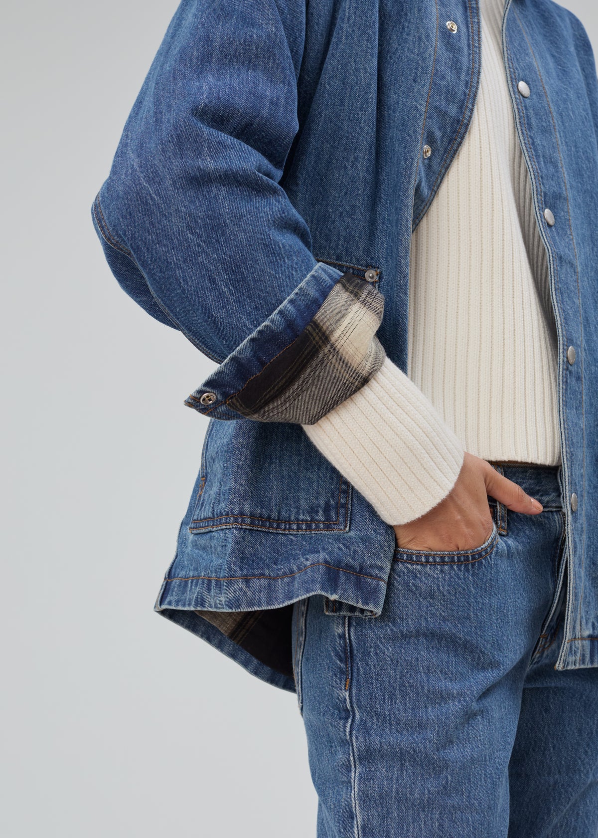 Barn Jacket in Classic Blue made of cotton denim with a rolled cuff revealing a plaid lining, worn over a cream ribbed sweater and blue jeans.