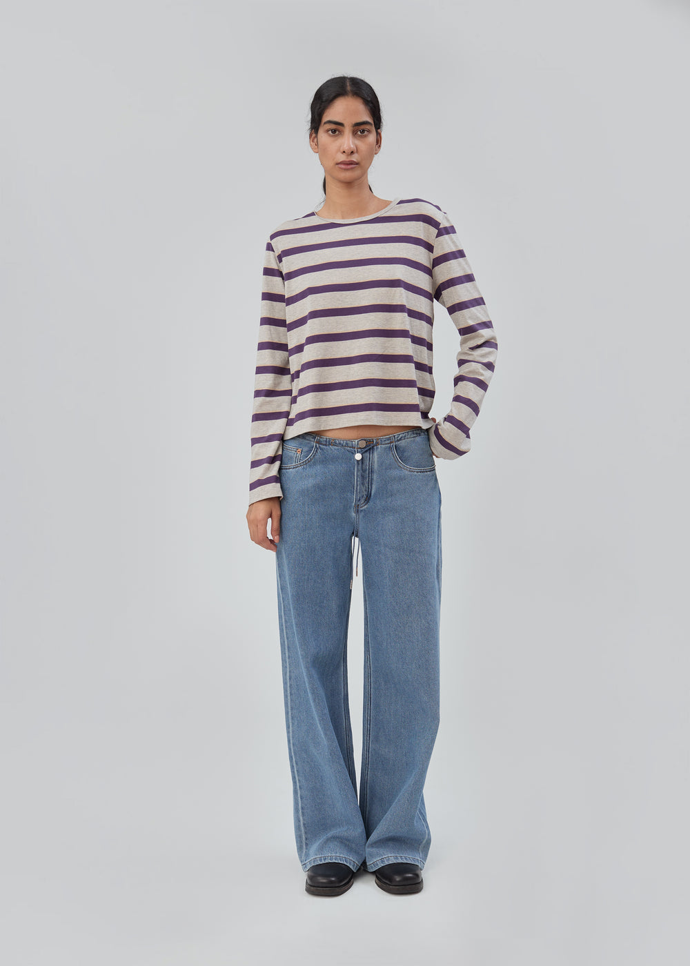 A person stands against a plain background wearing a long-sleeve purple and beige striped shirt, black shoes, and the Cool Jean in Classic Blue with a relaxed leg in the softest denim.