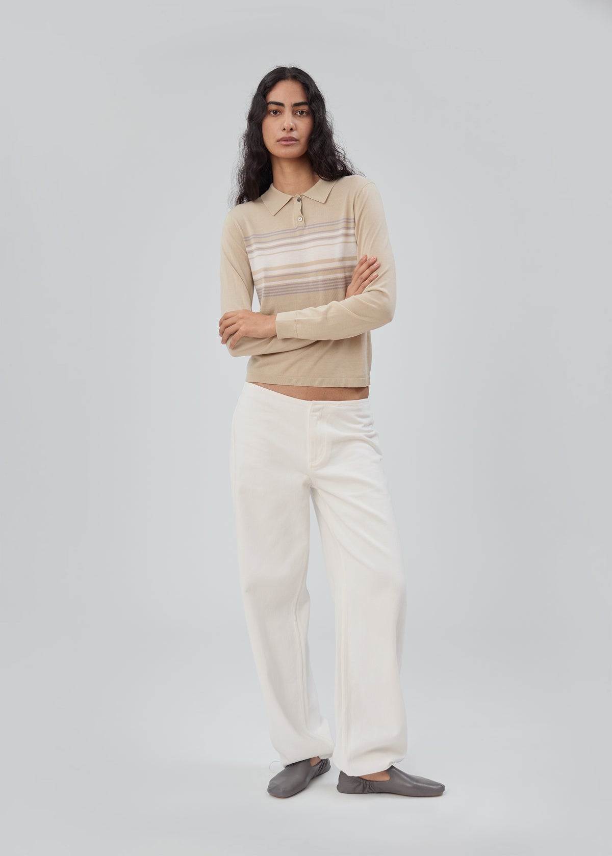 A model wears a beige long-sleeve Daughter Sweater Long Sleeve in Beige Stripe with horizontal stripes across the chest, paired with wide-leg white trousers and gray flats.