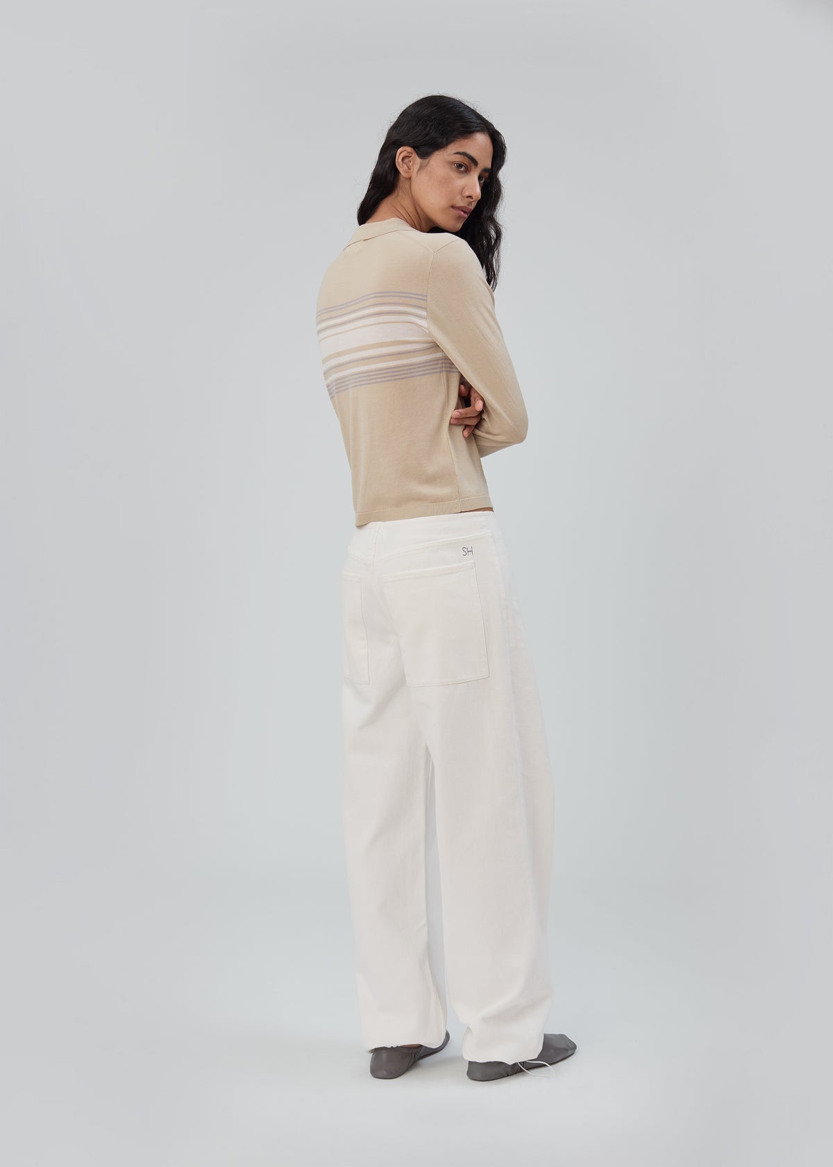 A person wearing a beige, long-sleeved, lightweight Daughter Sweater Long Sleeve in Beige Stripe with horizontal stripes across the back and wide-leg white pants.
