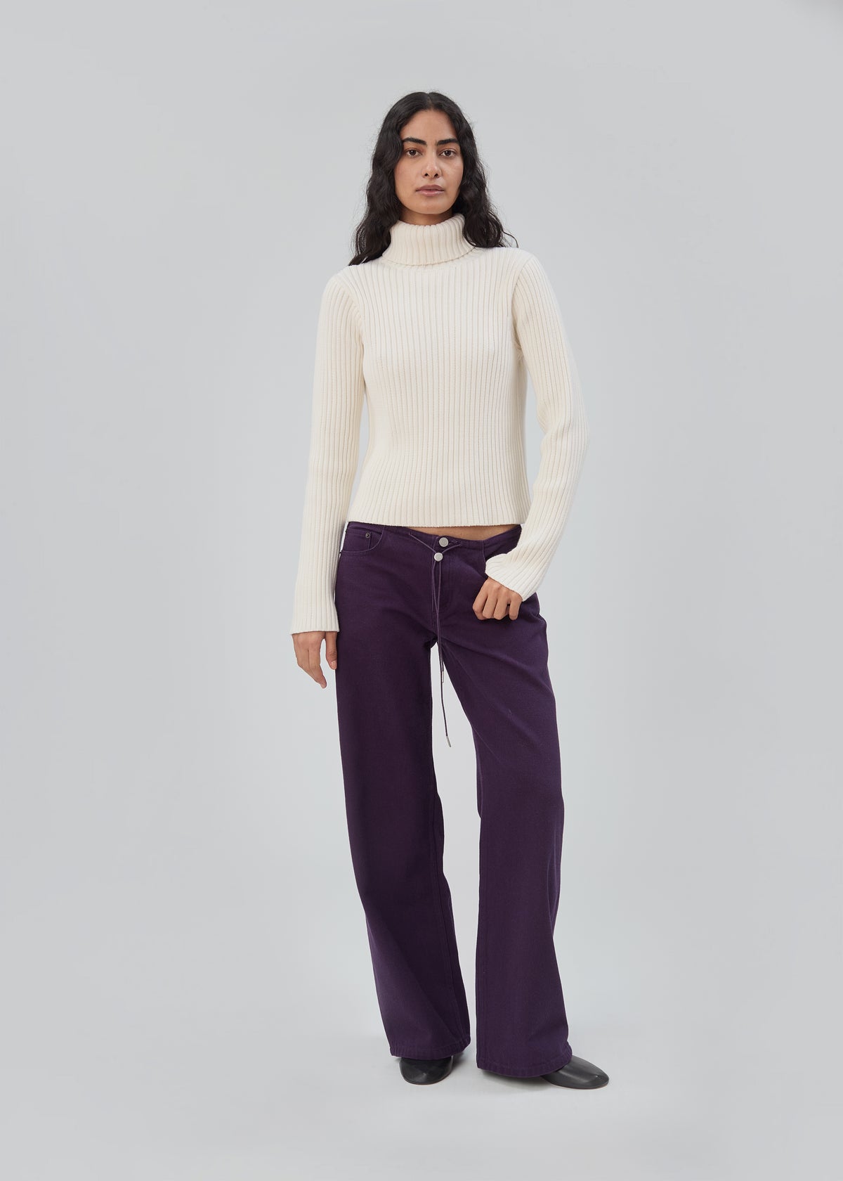 A model wears a cream turtleneck sweater and purple Cool Jean in Purple, which are low-rise, relaxed-leg jeans.
