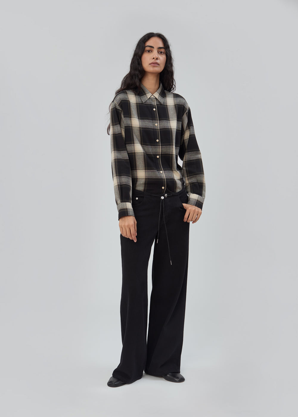 A person wearing a black and grey plaid button-down shirt tucked into Cool Jean in Black Black, featuring a relaxed leg and a thin drawstring belt.