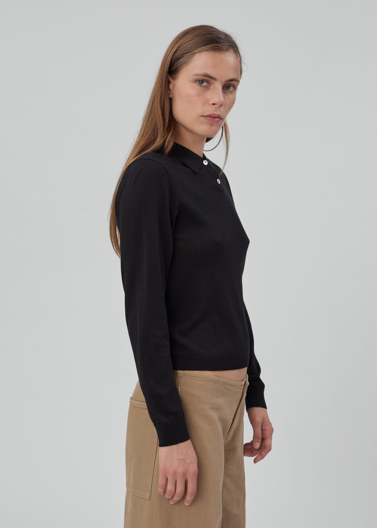 A woman wearing a black Daughter Sweater Long Sleeve in a knit polo style with a button placket and tan trousers.
