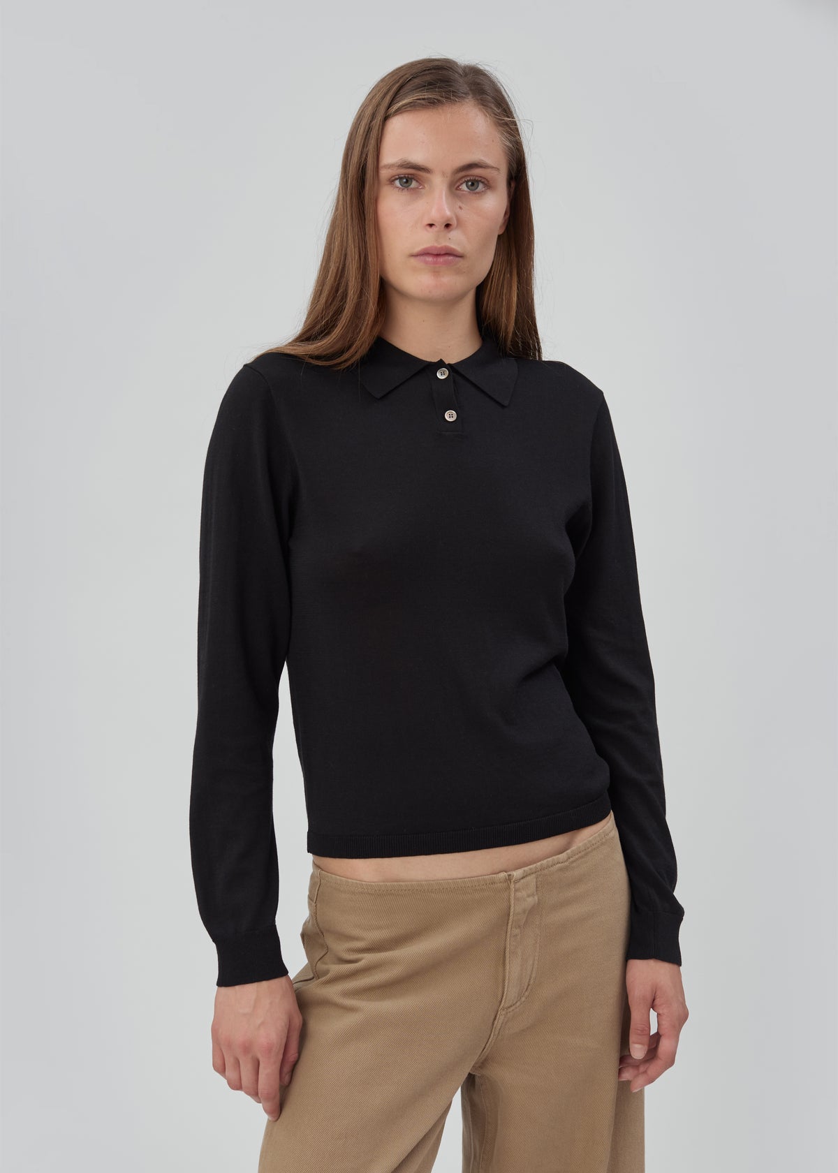 A person wearing a black Daughter Sweater Long Sleeve in a lightweight knit with a button-down collar and tan trousers.