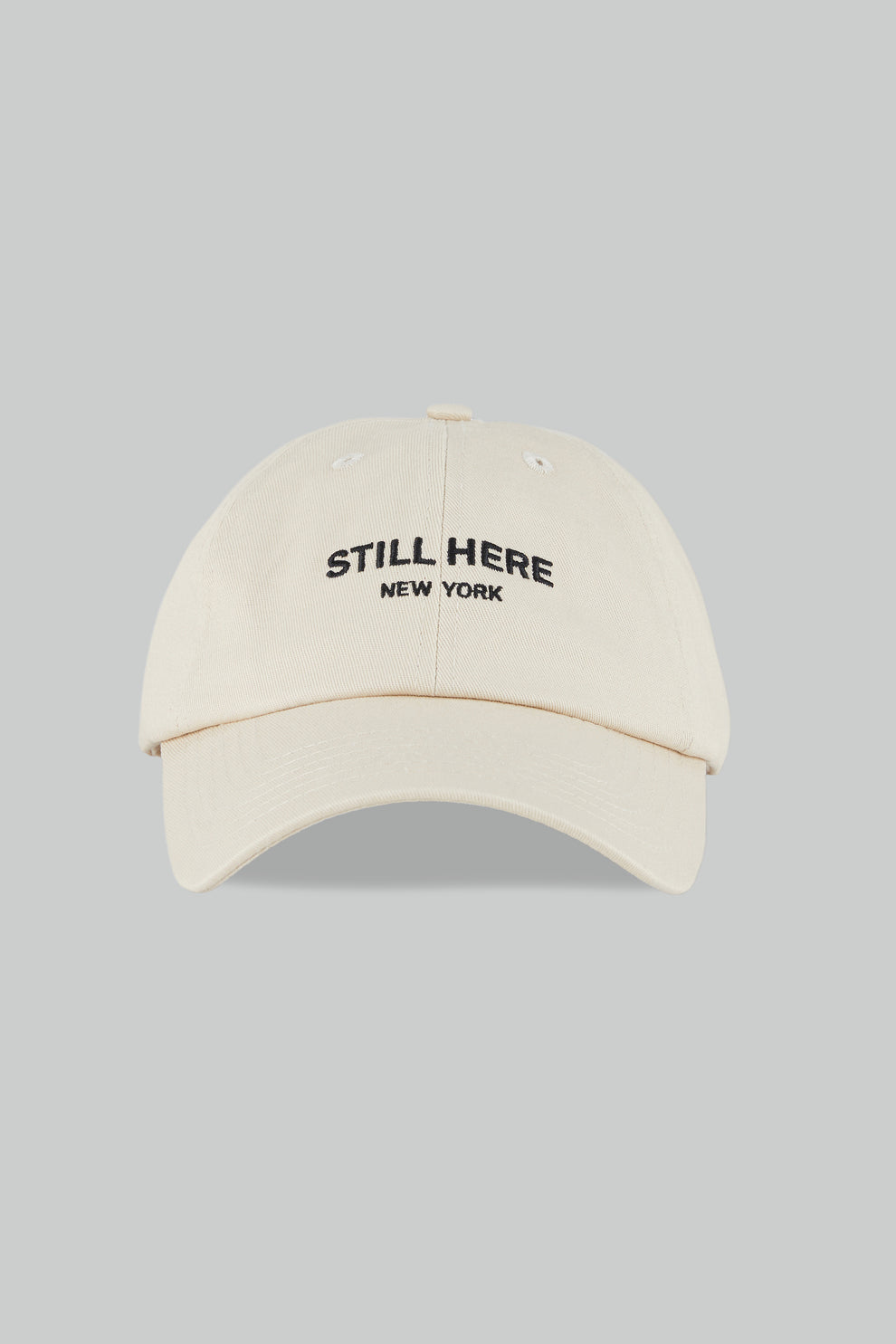 Beige cap with 'STILL HERE NEW YORK' text on a gray background
