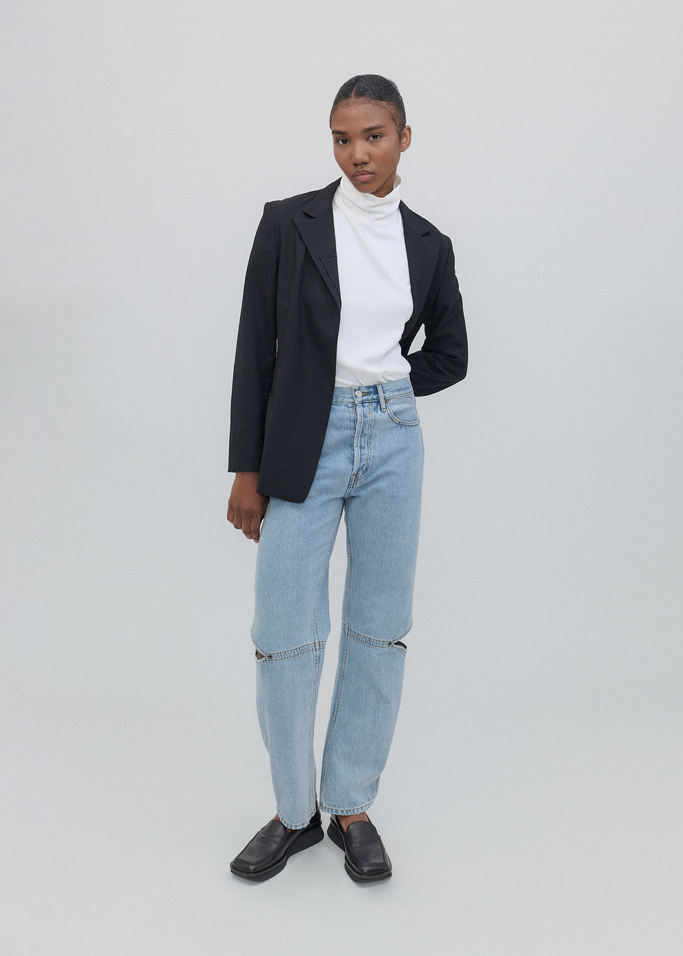 Person wearing a black blazer, white shirt, and light blue jeans on a plain background