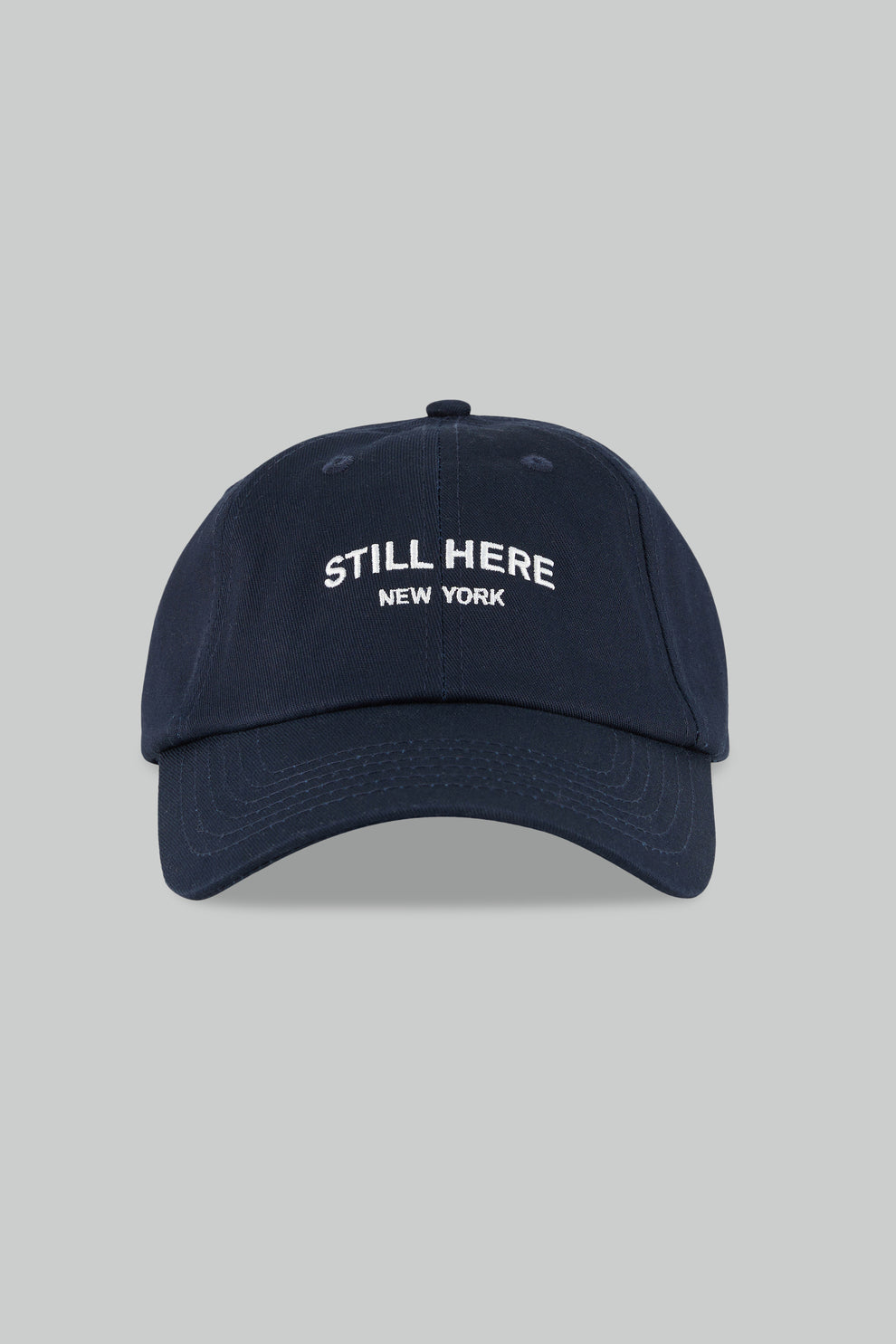 Navy blue cap with 'STILL HERE NEW YORK' text on a gray background