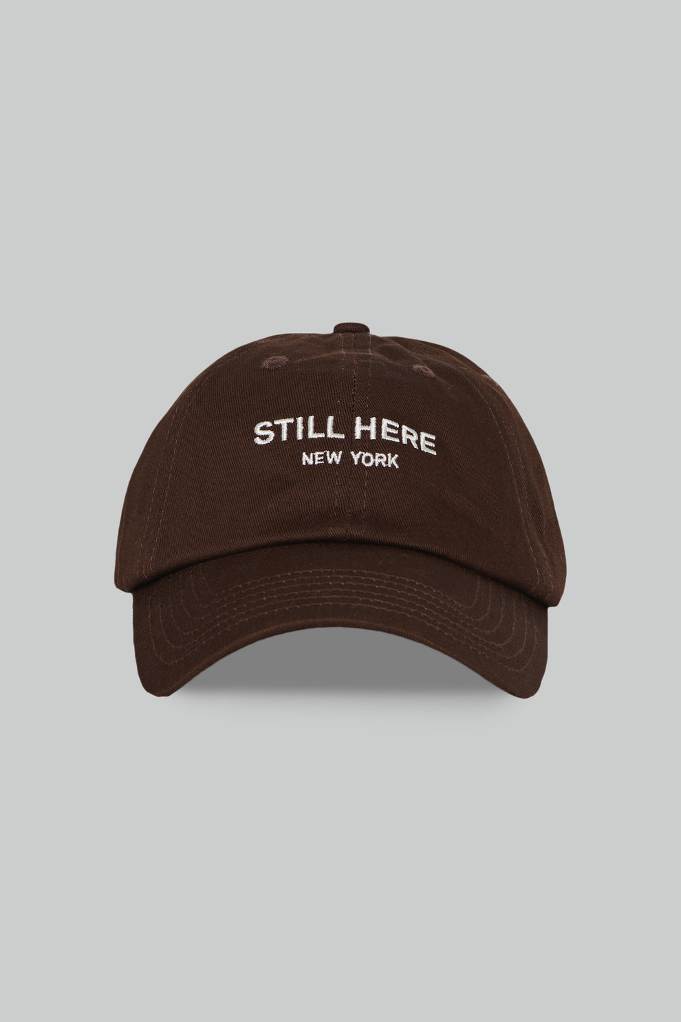 Brown cap with 'STILL HERE NEW YORK' text on a gray background